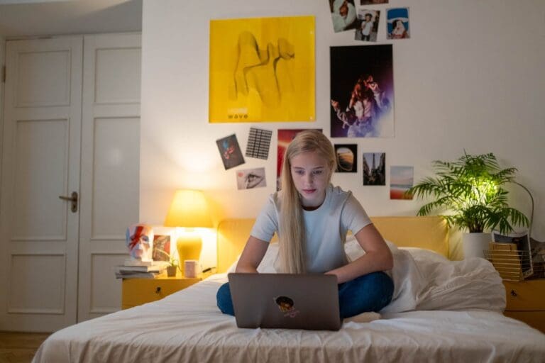 A girl sits cross-legged on a bed using a laptop in a bedroom with posters, a plant, and a lit lamp on a nightstand, perhaps engaging in an online therapy session with her therapist NYC or exploring Brooklyn Therapy resources from the comfort of her room.