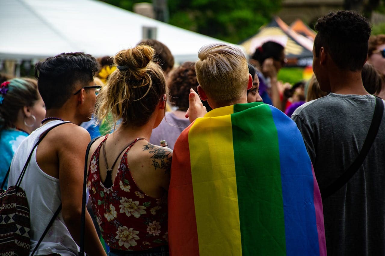 A group of people seen from behind, one person wearing a rainbow pride flag, standing outdoors at a crowded event—an atmosphere that reflects the vibrant inclusivity often celebrated in Brooklyn therapy communities and by supportive therapist NYC professionals.