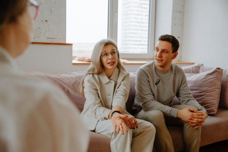 Three people sit on a couch in a bright room, engaged in what appears to be a Brooklyn Therapy session, with one woman speaking and gesturing while the others listen attentively. A window and light-colored pillows are visible in the background.