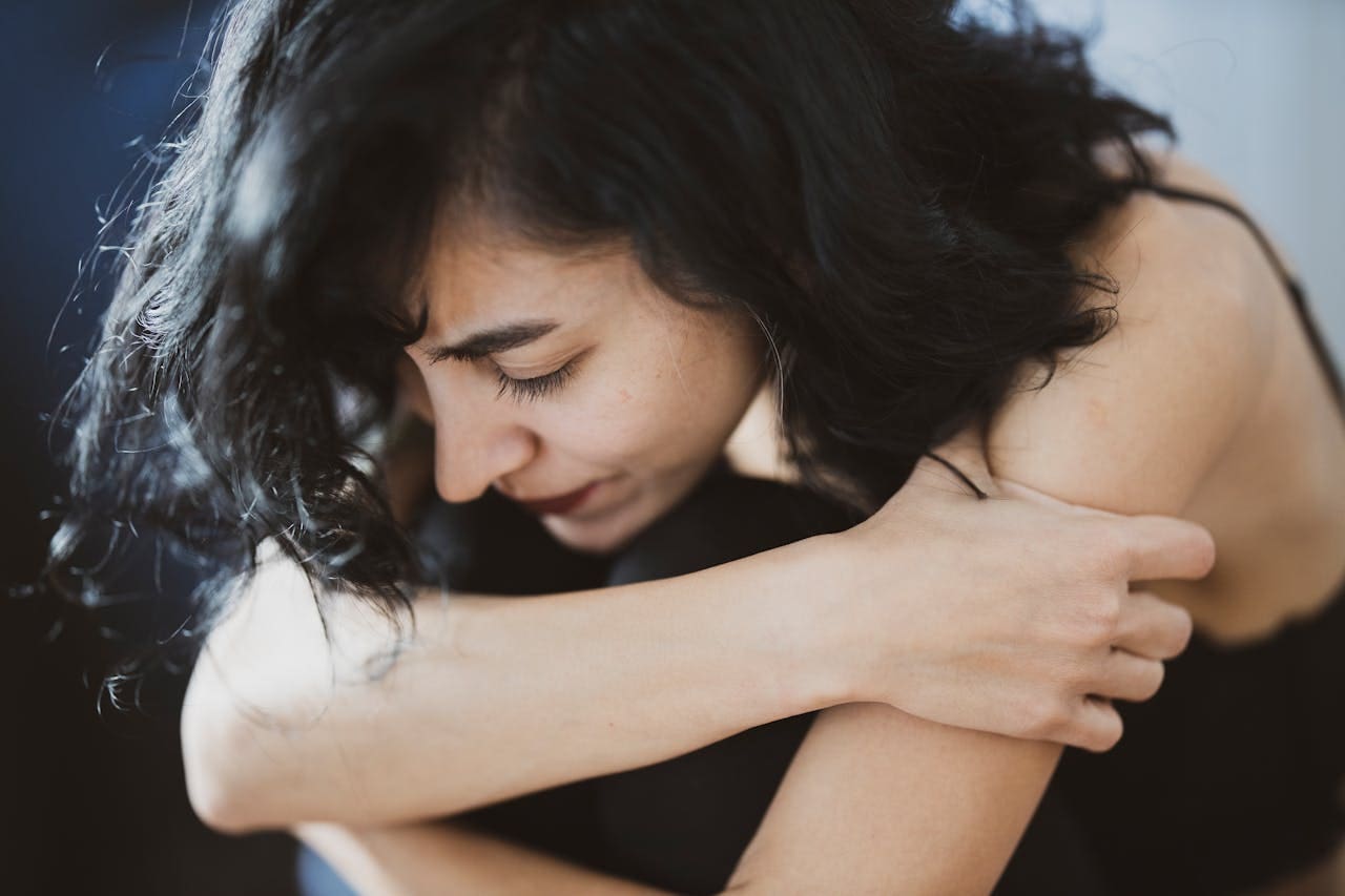 Woman with dark hair sits and hugs her knees, looking down with a distressed expression, perhaps contemplating whether Brooklyn Therapy or a trusted therapist in NYC could help her find comfort and support.