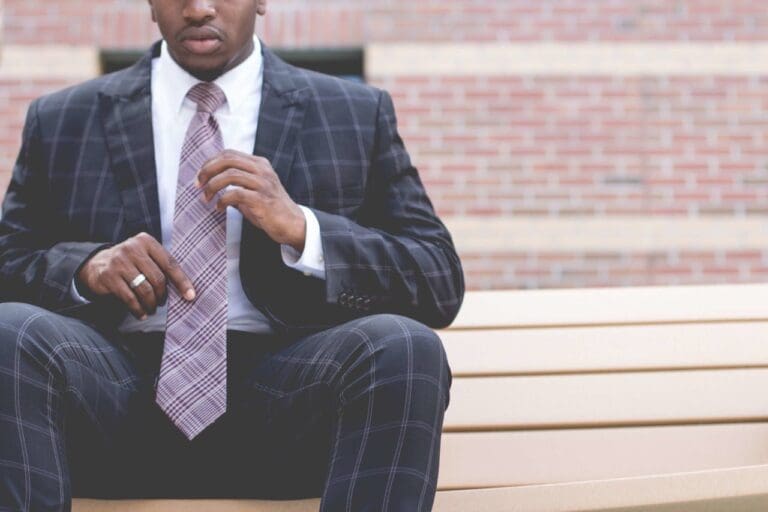 A man in a plaid suit and tie sits on a bench, adjusting his tie—perhaps preparing for his next therapy session. A brick wall is visible in the background, evoking the urban charm of Brooklyn Therapy.