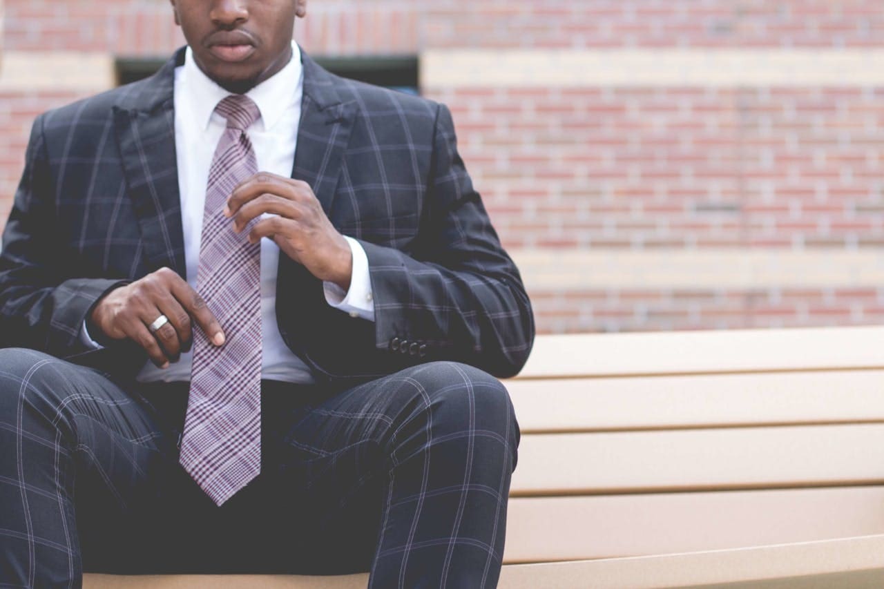 A man in a plaid suit and tie sits on a bench, adjusting his tie—perhaps preparing for his next therapy session. A brick wall is visible in the background, evoking the urban charm of Brooklyn Therapy.
