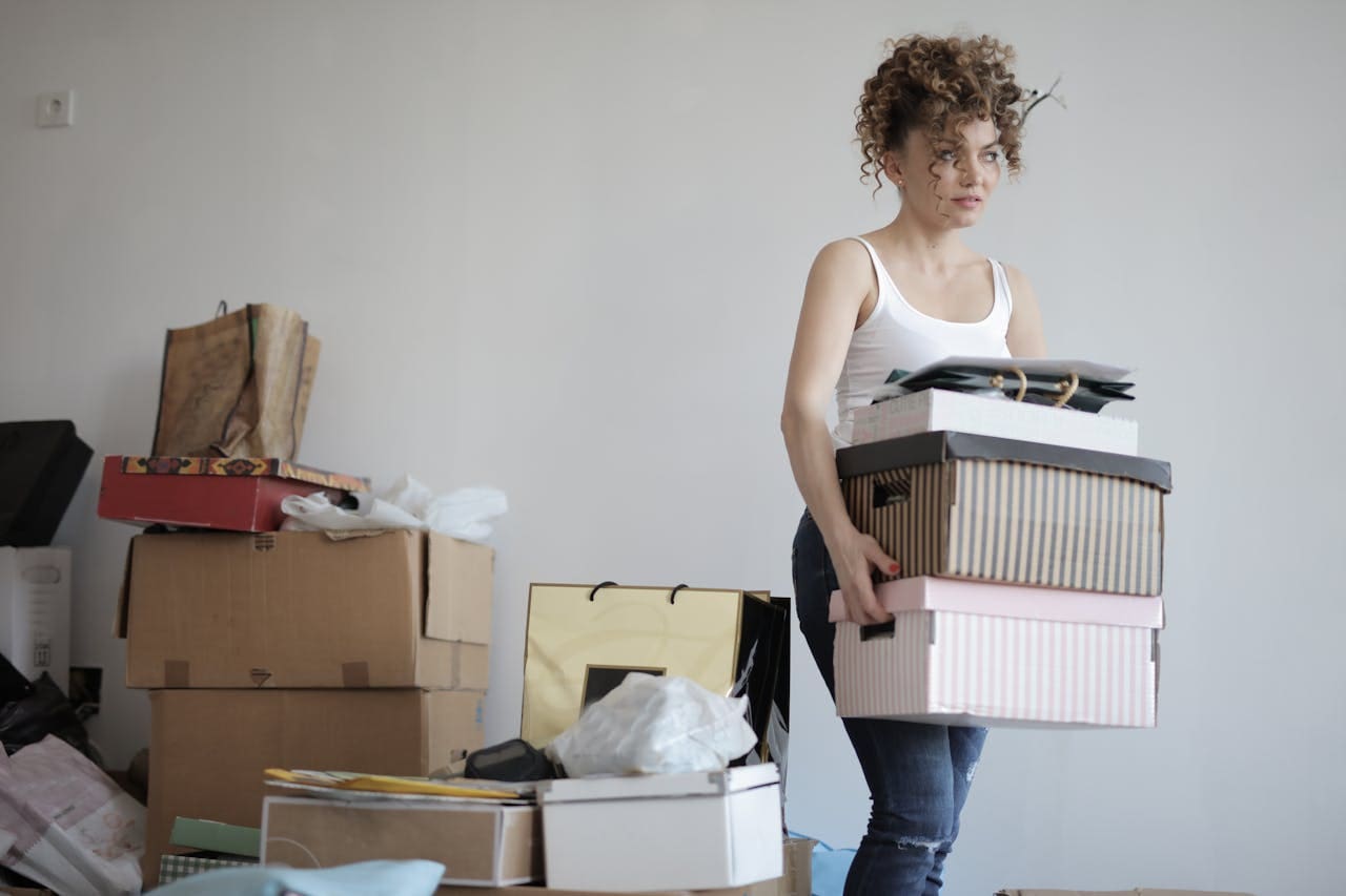 A woman in a white tank top carries stacked boxes in a room filled with moving boxes and bags, reflecting the fresh start that often comes with change—and sometimes the support of Brooklyn Therapy or a trusted therapist NYC.