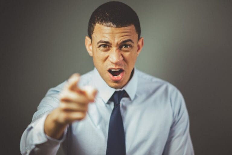 Man in a light blue shirt and dark tie pointing directly at the camera with an intense, open-mouthed expression against a plain background, capturing the energy and directness often seen in Brooklyn therapy sessions or when meeting a therapist NYC clients trust.