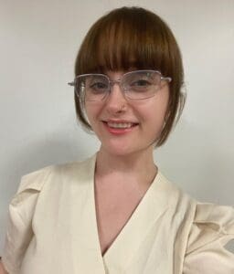 A person with short brown hair, bangs, clear glasses, and a nose ring smiles at the camera. They are wearing a cream-colored top against a plain light background, capturing the welcoming spirit often found in Brooklyn Therapy settings.