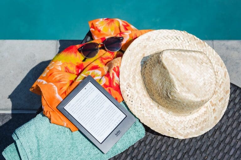 A straw hat, sunglasses, an orange floral shirt, a teal towel, and an e-reader are placed beside a swimming pool on a lounge chair—setting the perfect scene for relaxation after a Brooklyn Therapy session.