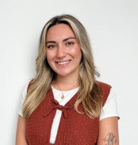 A woman with long blonde hair wearing a white shirt and a rust-colored sweater vest stands against a white background, smiling at the camera—radiating the warmth and professionalism you’d expect from a trusted Brooklyn Therapy provider.