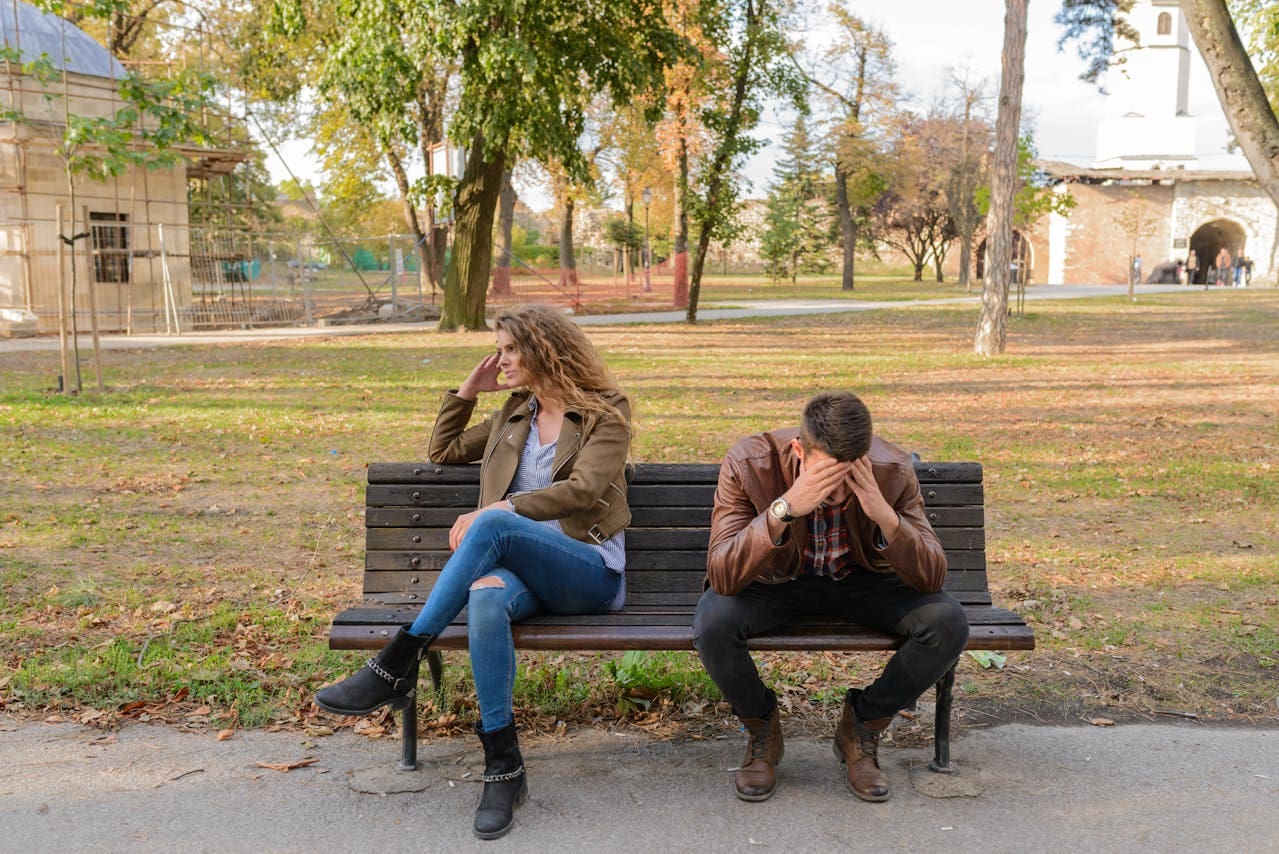A woman and man sit on a park bench, both looking upset. The woman faces away with her hand on her head, while the man leans forward, covering his face with his hands—perhaps considering whether therapy or speaking to a therapist in NYC could help them move forward.