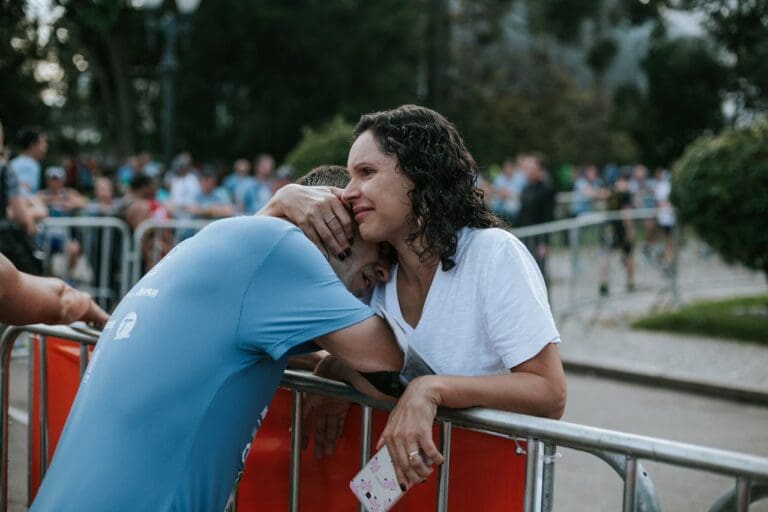 A woman in a white shirt hugs a man in a blue shirt over a barrier at an outdoor event, with a crowd and trees in the background—a heartfelt moment that reflects the support and connection valued by Brooklyn Therapy.