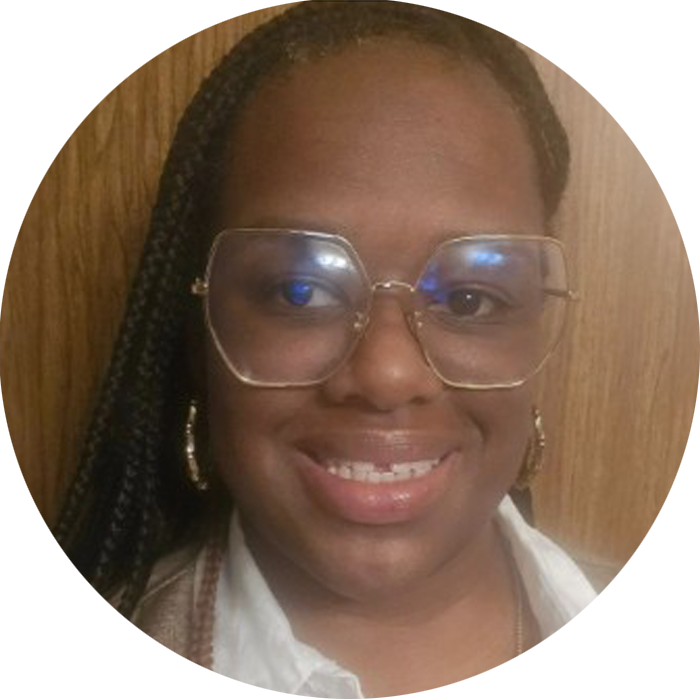 A person with long braided hair, large square glasses, and gold earrings smiles in front of a wooden background, radiating warmth and approachability—perfect qualities for someone seeking Brooklyn Therapy or searching for a compassionate therapist in NYC.