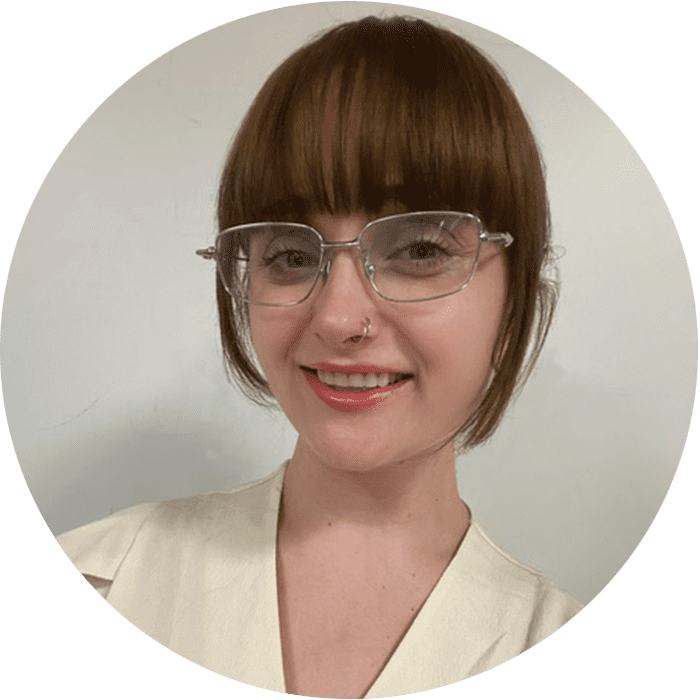 A person with short brown hair, bangs, glasses, and a nose ring is smiling at the camera. They are wearing a light-colored top, radiating warmth and approachability—qualities that are often found in a great therapist NYC residents trust for their therapy needs.