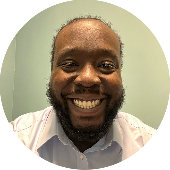 A man with a beard and mustache smiles at the camera, wearing a light-colored collared shirt against a pale green background—portraying the calm confidence you might find with a therapist NYC trusts at Brooklyn Therapy.