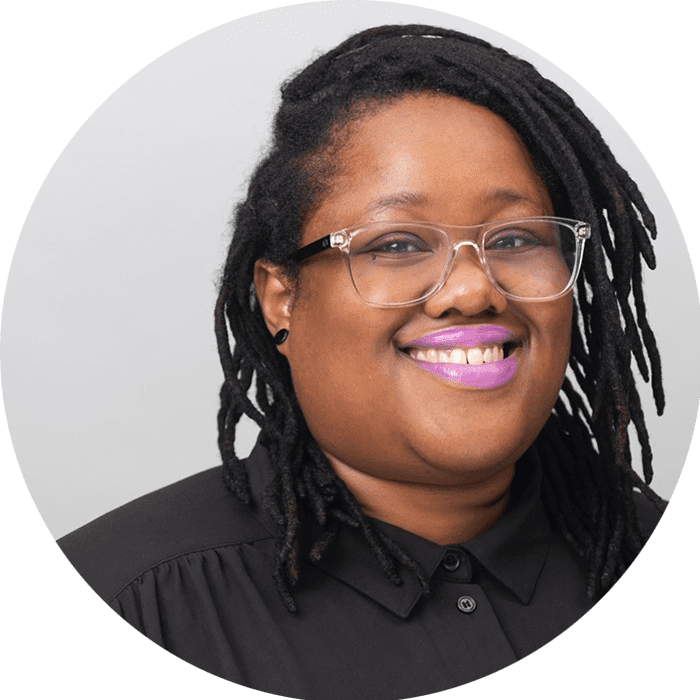 A person with glasses and long, black locs smiles at the camera, wearing a black button-up shirt and purple lipstick against a light background—capturing the approachable presence you’d expect from a Brooklyn Therapy professional or therapist NYC clients trust.
