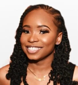 A woman with long, twisted hair smiles at the camera. She is wearing a gold necklace and earrings, with a neutral background behind her—a warm, inviting image often seen on Brooklyn Therapy and therapist NYC profiles.
