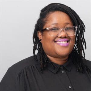 Person with glasses and locs, wearing a black button-up shirt, smiles against a plain light gray background—capturing the warm and welcoming spirit of Brooklyn Therapy for those seeking compassionate therapy in NYC.