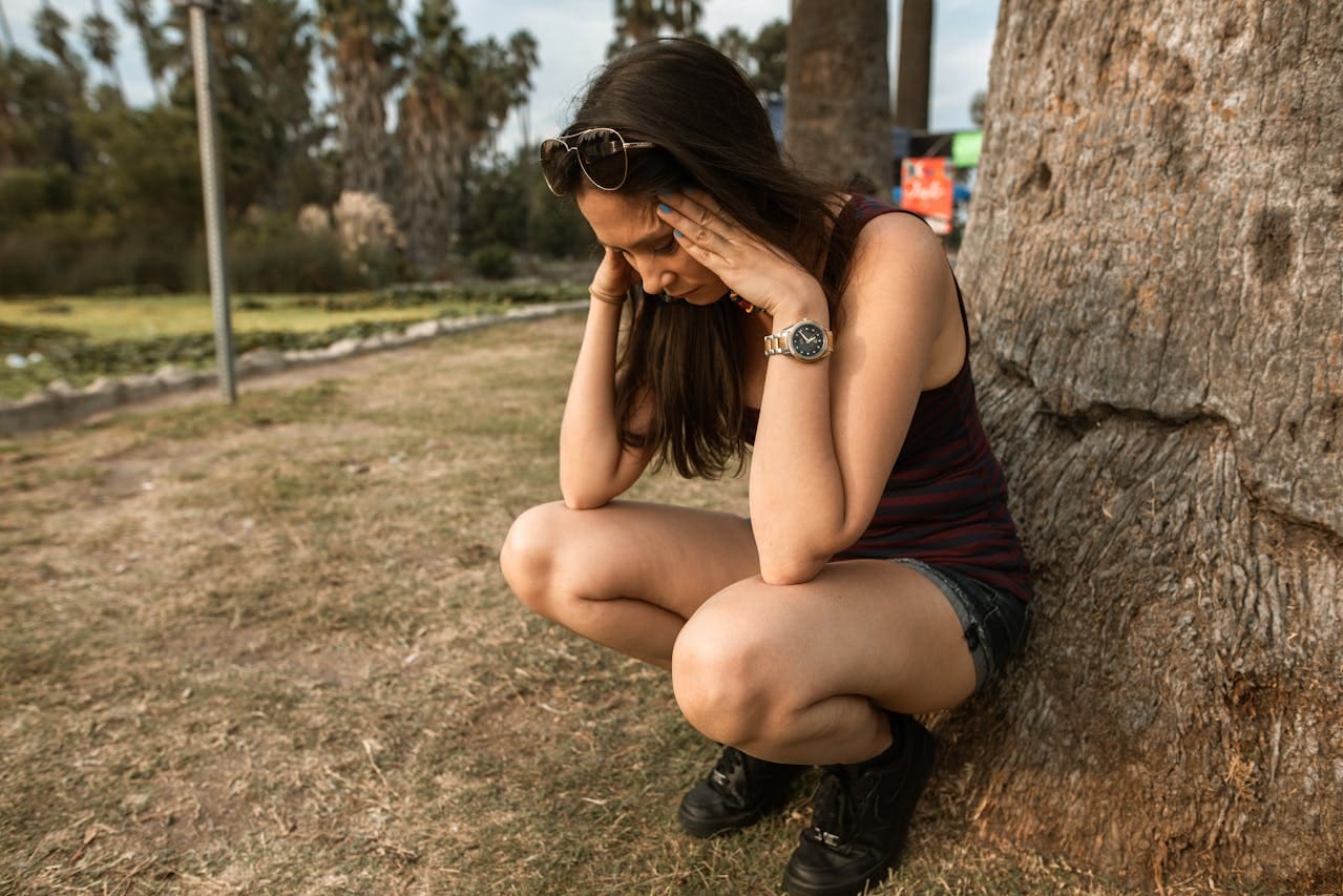 Woman squatting outside by a tree, holding her head in her hands, wearing sunglasses, a striped tank top, shorts, and boots, appearing stressed or deep in thought—perhaps she’s considering reaching out for Brooklyn Therapy to find support or speak with a therapist NYC locals trust.