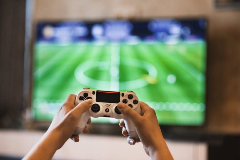 Person holding a white video game controller in front of a large TV displaying a soccer video game, illustrating how some people use gaming as a form of relaxation or stress relief—something often discussed in Brooklyn Therapy sessions.