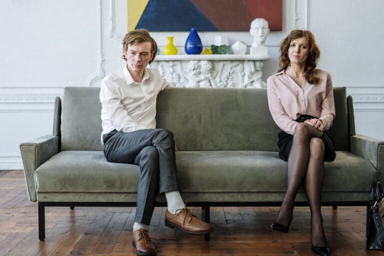 A man and a woman sit apart on a gray couch, looking away from each other in a bright room with a fireplace and modern art—capturing the quiet tension often explored in Brooklyn Therapy sessions.