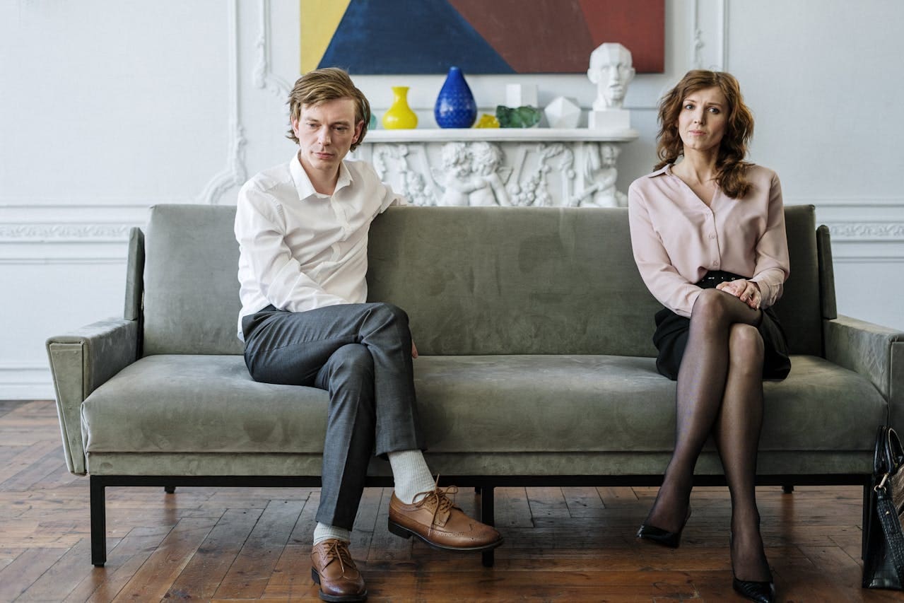 A man and a woman sit apart on a gray couch, looking away from each other in a bright room with a fireplace and modern art—capturing the quiet tension often explored in Brooklyn Therapy sessions.