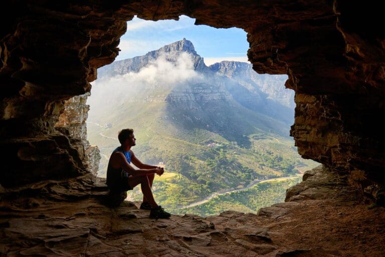 A person sits at the entrance of a rocky cave, overlooking a green valley and distant mountains partially covered by clouds under a blue sky—a peaceful scene that evokes the serenity often sought in therapy.