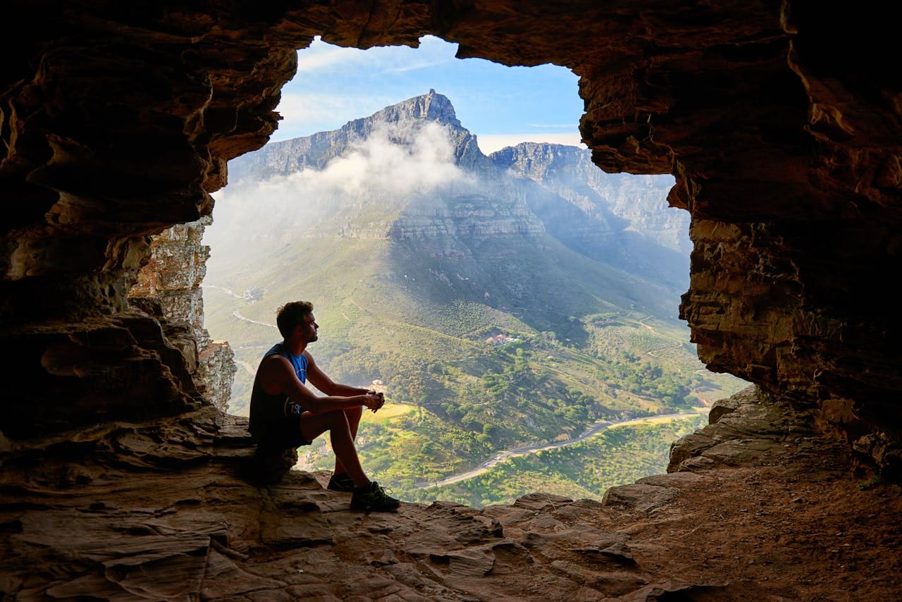 A person sits at the entrance of a rocky cave, overlooking a green valley and distant mountains partially covered by clouds under a blue sky—a peaceful scene that evokes the serenity often sought in therapy.