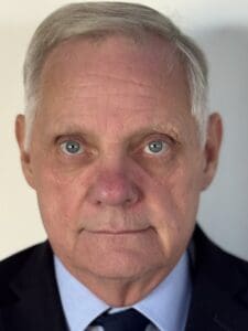 Older man with short gray hair in a suit and tie, staring directly at the camera with a neutral expression against a plain background—an image that reflects the calm professionalism often found in experienced Brooklyn Therapy practitioners and therapist NYC offices.