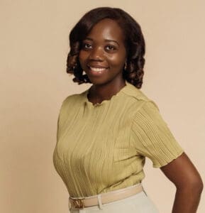 A woman with shoulder-length curled hair, wearing a light green pleated blouse and beige pants, stands smiling against a plain beige background—conveying warmth and approachability often sought in a therapist NYC or Brooklyn Therapy setting.