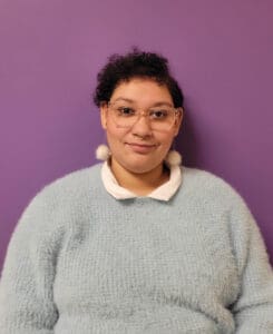 A person with short curly hair, wearing glasses, a light blue fuzzy sweater, white collared shirt, and pom-pom earrings sits in front of a solid purple background—capturing the warm, approachable vibe often found in Brooklyn Therapy settings.