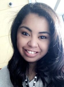 A woman with long dark hair smiles at the camera indoors, wearing a gray top and a beaded necklace. A security camera is visible on the wall behind her, hinting at a safe and welcoming space—perhaps reflective of a Brooklyn Therapy office or the comfortable environment provided by a trusted therapist NYC.