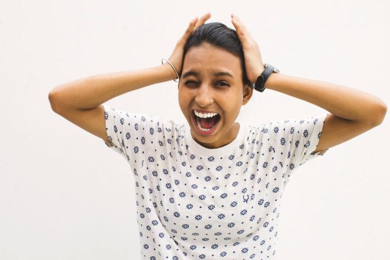 Woman in a patterned shirt stands against a plain white background, holding her head with both hands and shouting with her mouth open—as if expressing the frustration that often leads people to seek therapy.