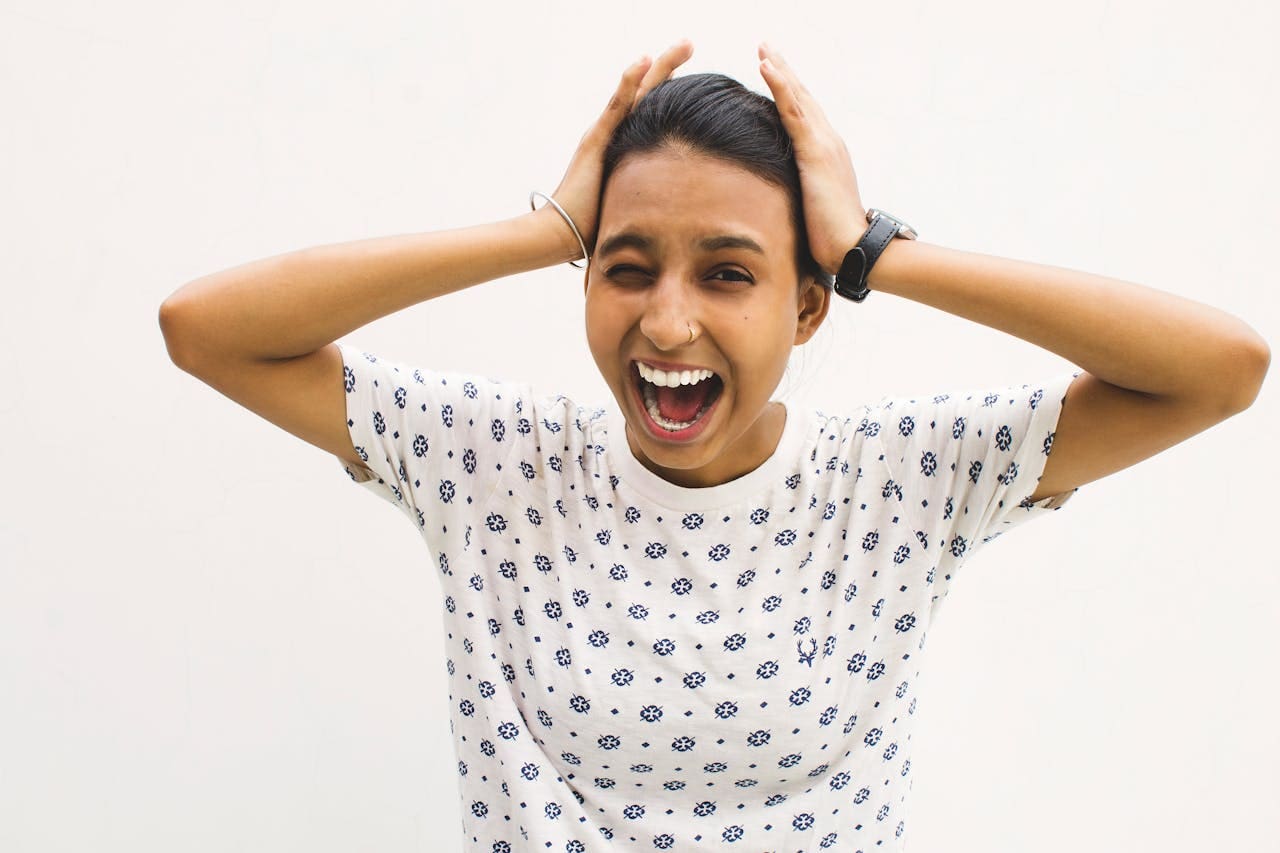 Woman in a patterned shirt stands against a plain white background, holding her head with both hands and shouting with her mouth open—as if expressing the frustration that often leads people to seek therapy.