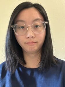 Person with straight black hair and clear glasses wearing a navy blue shirt, looking at the camera against a plain light-colored background, embodying the calm and approachable presence you might find in a Brooklyn Therapy office or with a therapist NYC clients trust.