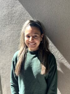 A person with long dark hair wearing a green sweatshirt stands against a textured wall, smiling in bright sunlight with a diagonal shadow behind them—capturing the warmth and approachability often found in Brooklyn Therapy sessions.