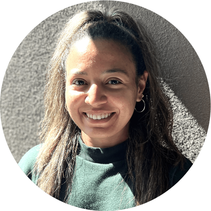 Woman with long brown hair wearing a green sweater and hoop earrings, smiling in front of a textured gray wall with sunlight casting a shadow—a warm and welcoming presence, perfect for someone offering Brooklyn Therapy.