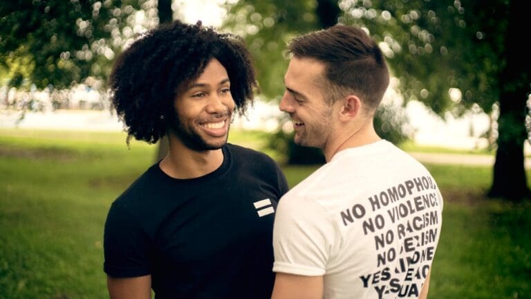 Two men stand facing each other and smiling in a park. One wears a black shirt; the other wears a white shirt with anti-discrimination messages printed on the back, creating a moment that reflects the supportive spirit of Brooklyn Therapy and highlights the power of connection found through therapy in NYC.