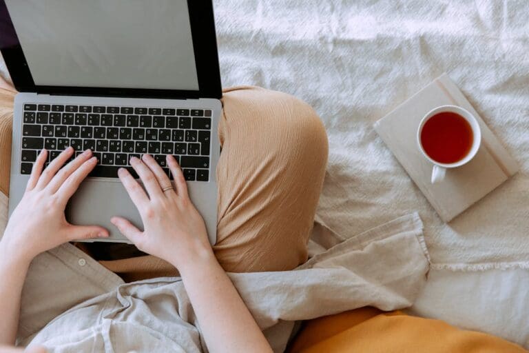 Person typing on a laptop while sitting on a bed, with a cup of tea or coffee placed on a closed book nearby—an inviting scene that reflects the comfort and introspection often found in Brooklyn Therapy sessions or while connecting with a therapist NYC remotely.