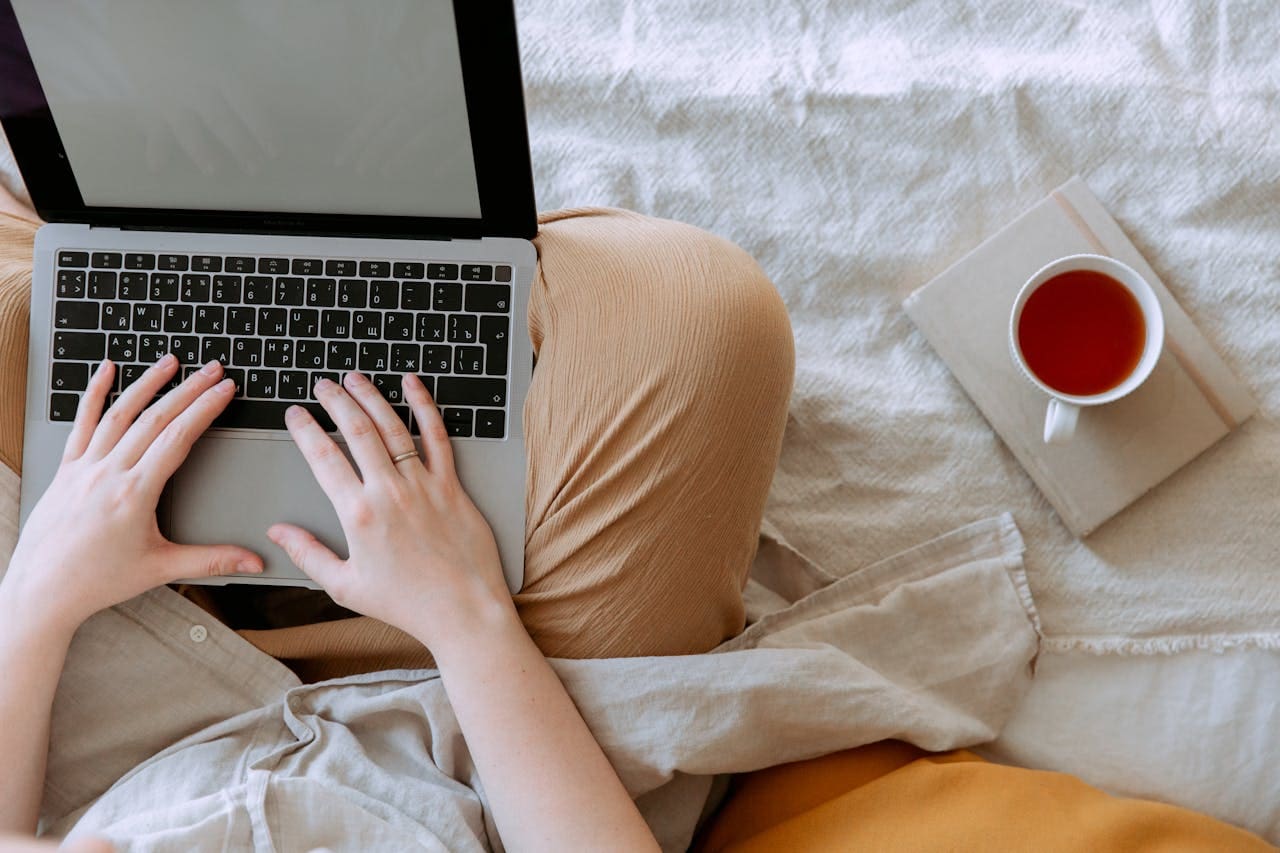 Person typing on a laptop while sitting on a bed, with a cup of tea or coffee placed on a closed book nearby—an inviting scene that reflects the comfort and introspection often found in Brooklyn Therapy sessions or while connecting with a therapist NYC remotely.