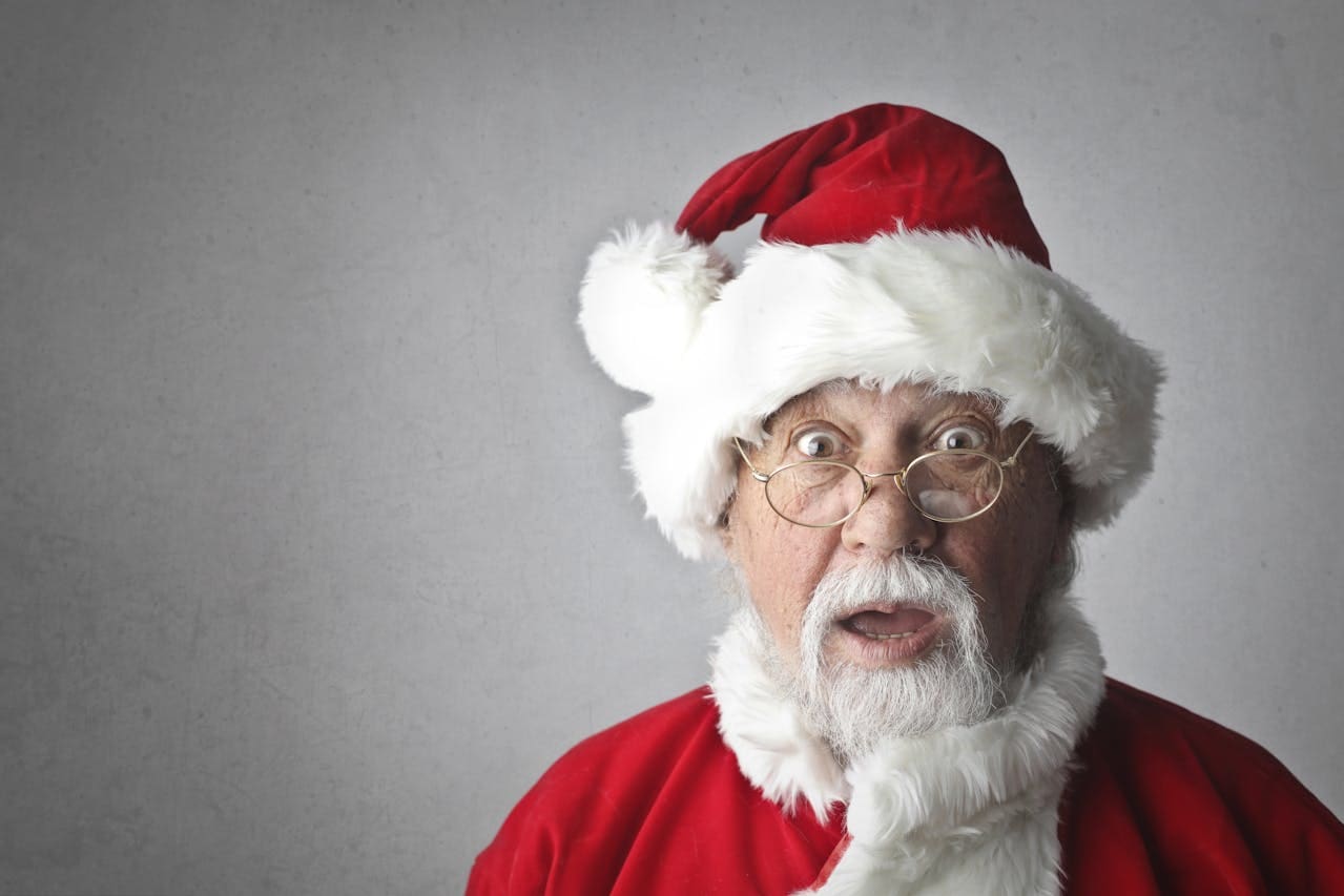 Older adult with a white beard wearing a red Santa hat and suit, looking surprised, standing in front of a plain light background—perfect for promoting Brooklyn Therapy during the holiday season.