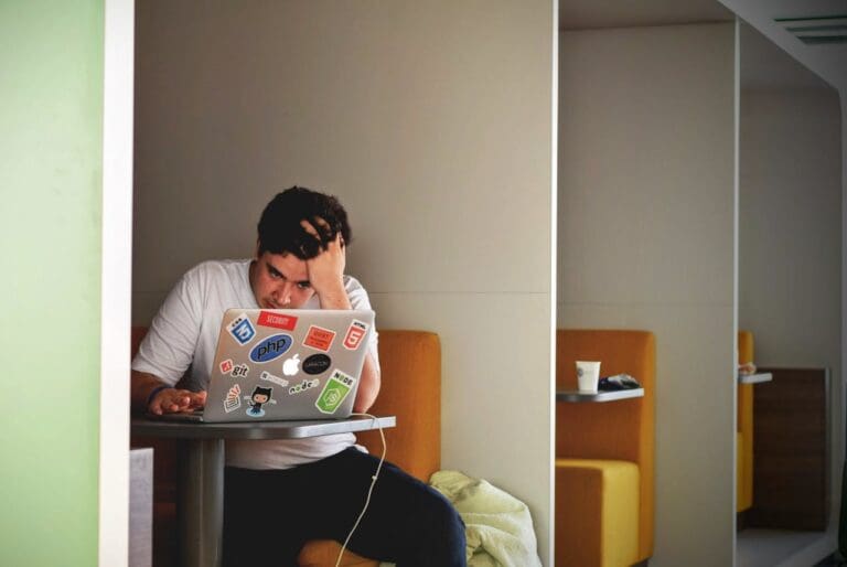 A person sits alone in a booth, looking stressed while using a laptop covered in programming stickers—a scene that highlights the need for support such as Brooklyn Therapy. A cup and jacket are on the table.