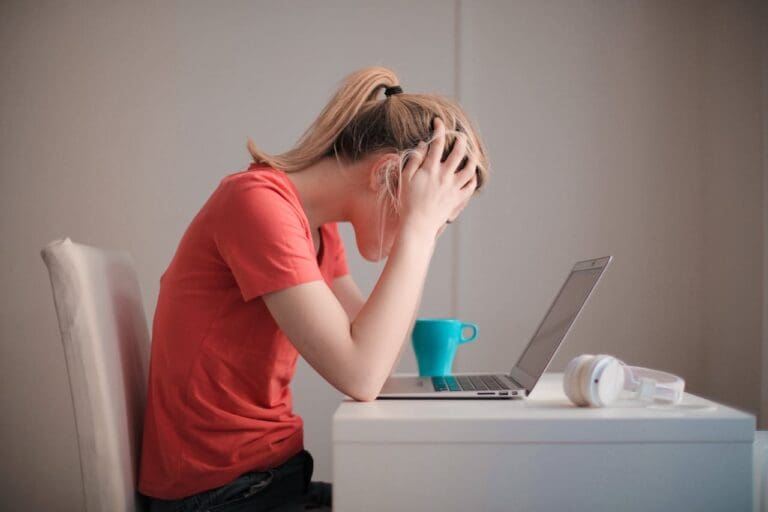 A person in a red shirt sits at a desk with a laptop, holding their head in their hands, appearing stressed—a scene that highlights the need for support such as Brooklyn Therapy. A blue mug and white headphones are on the desk.