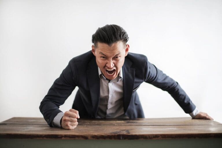 A man in a suit leans over a wooden table, shouting with a clenched fist, expressing anger or frustration against a plain white background—perhaps capturing the intense emotions often discussed during therapy sessions or explored in Brooklyn Therapy.