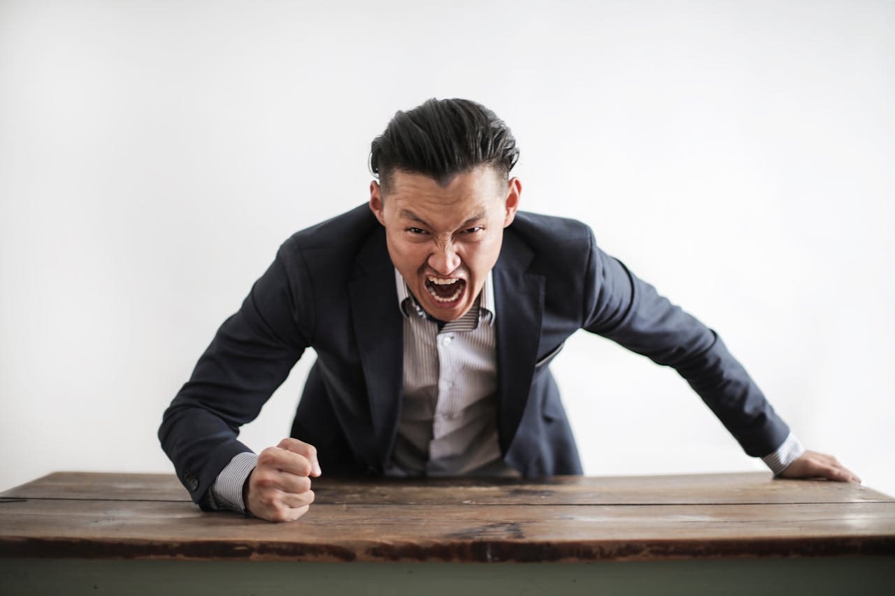 A man in a suit leans over a wooden table, shouting with a clenched fist, expressing anger or frustration against a plain white background—perhaps capturing the intense emotions often discussed during therapy sessions or explored in Brooklyn Therapy.