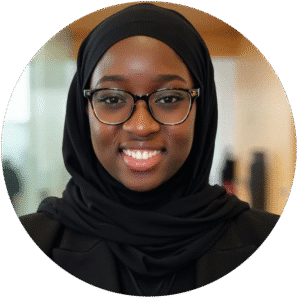 A woman wearing glasses and a black hijab smiles at the camera, standing indoors with a blurred background—an inviting presence that reflects the warmth and professionalism found in Brooklyn Therapy, one of the trusted therapy spaces in NYC.