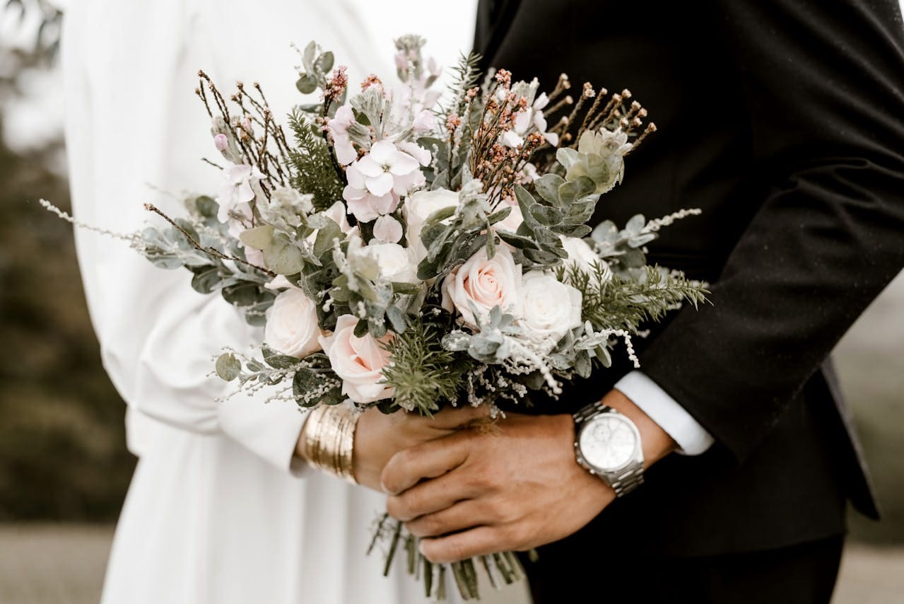A couple in formal attire holds a bouquet of white and pink flowers with greenery, standing close together outdoors—capturing a moment of connection that echoes the supportive relationships built through Brooklyn Therapy.