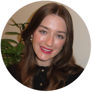 Woman with long brown hair and red lipstick smiling, wearing a black top with gold buttons, in front of a leafy green plant and a light-colored wall—capturing the warm, approachable vibe you’ll find at Brooklyn Therapy with an experienced therapist NYC residents trust.