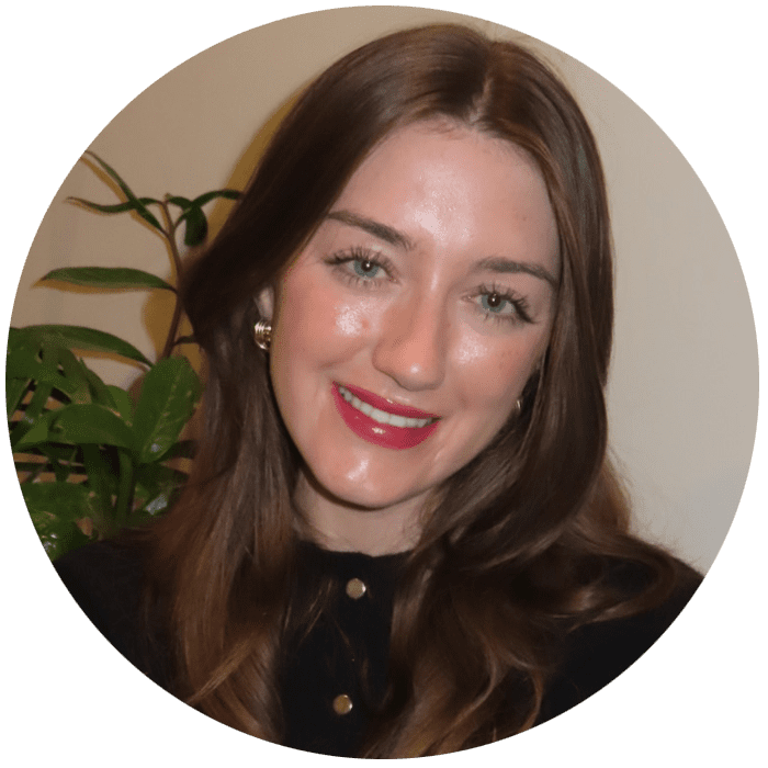 Woman with long brown hair and red lipstick smiling, wearing a black top with gold buttons, in front of a leafy green plant and a light-colored wall—capturing the warm, approachable vibe you’ll find at Brooklyn Therapy with an experienced therapist NYC residents trust.