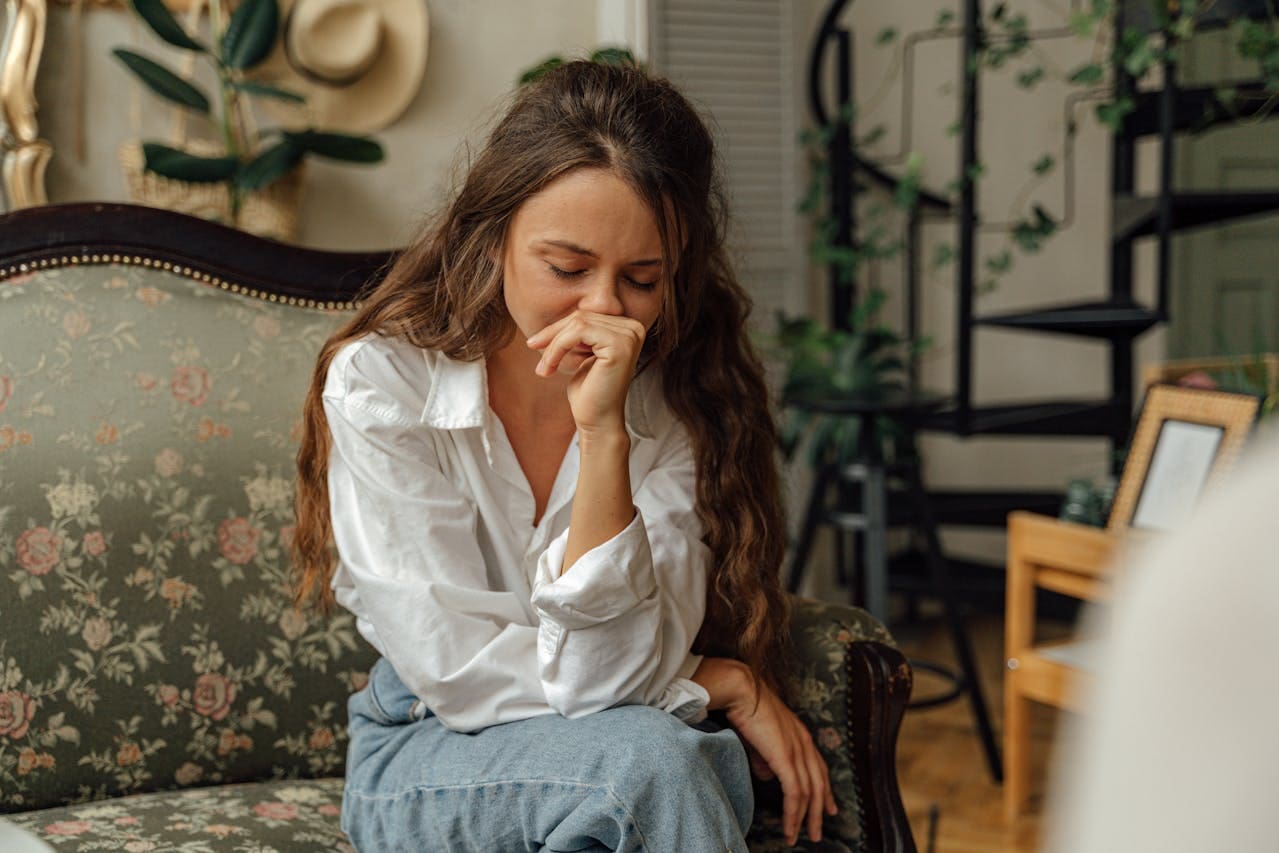 A woman with long brown hair sits on a sofa, resting her head on her hand and appearing deep in thought or upset, perhaps reflecting on recent sessions at Brooklyn Therapy with her therapist in NYC.