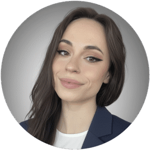 Woman with long brown hair, winged eyeliner, and a nose ring, wearing a blue blazer over a white top, poses in front of a neutral gray background—reflecting the calm professionalism often found in Brooklyn Therapy settings.
