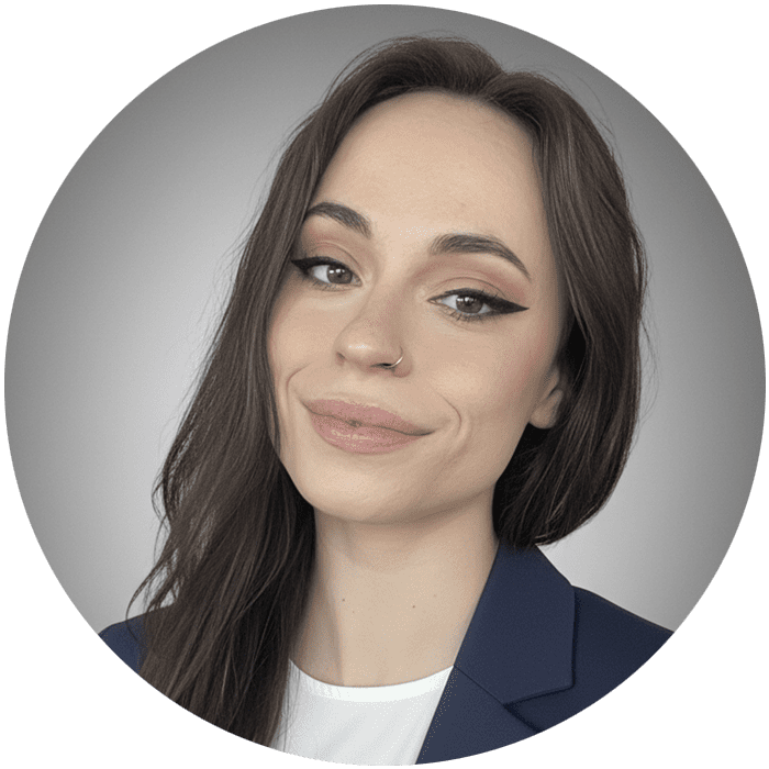 Woman with long brown hair, winged eyeliner, and a nose ring, wearing a blue blazer over a white top, poses in front of a neutral gray background—reflecting the calm professionalism often found in Brooklyn Therapy settings.