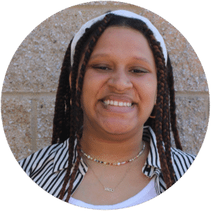 A woman with long braided hair, wearing a striped shirt and beaded necklace, smiles in front of a tan brick wall—embodying the warmth and approachability often found in Brooklyn Therapy sessions and therapy settings throughout NYC.